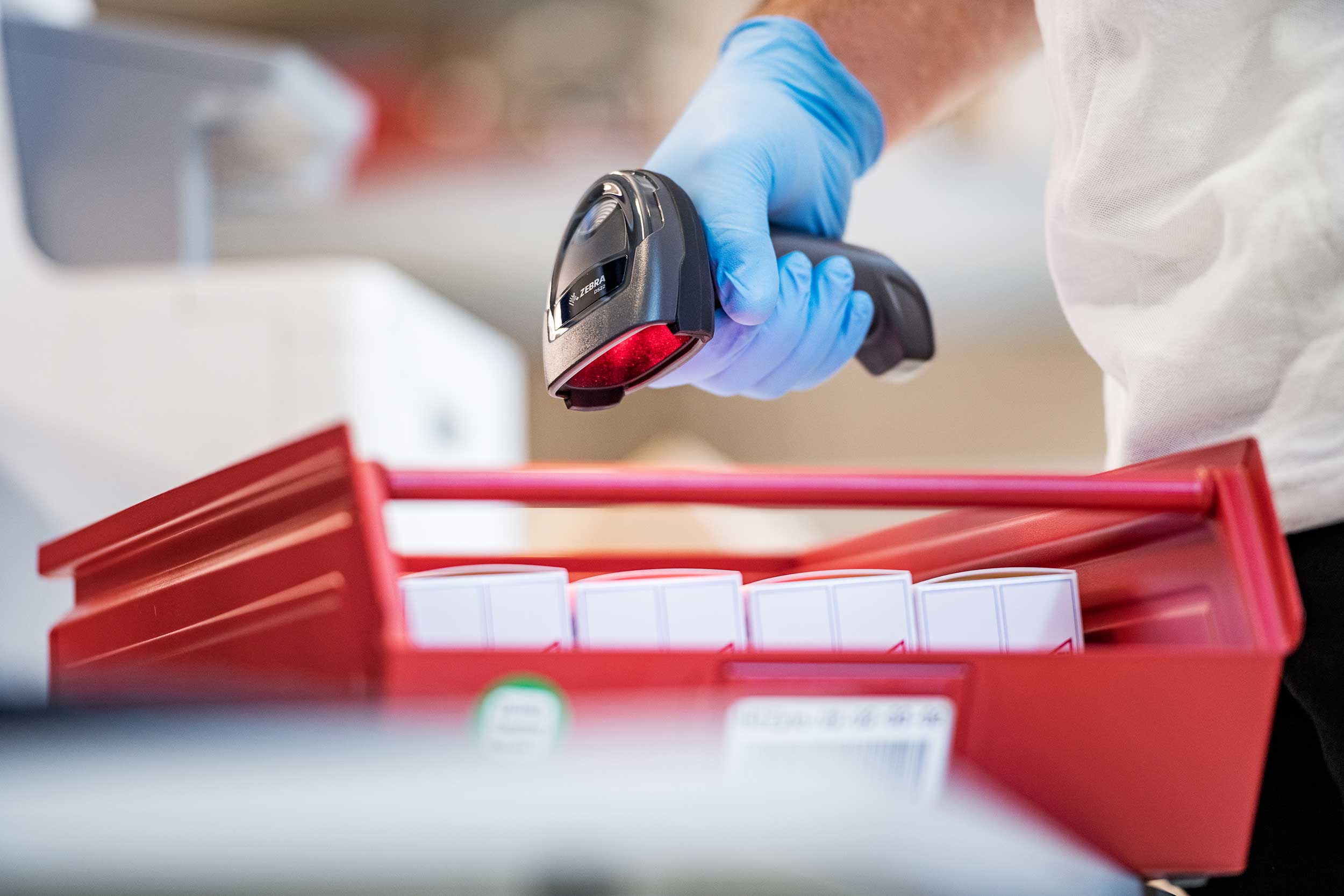Eine Person mit blauen Handschuhen scannt eine rote Box mit Medikamenten mithilfe eines Barcodescanners.