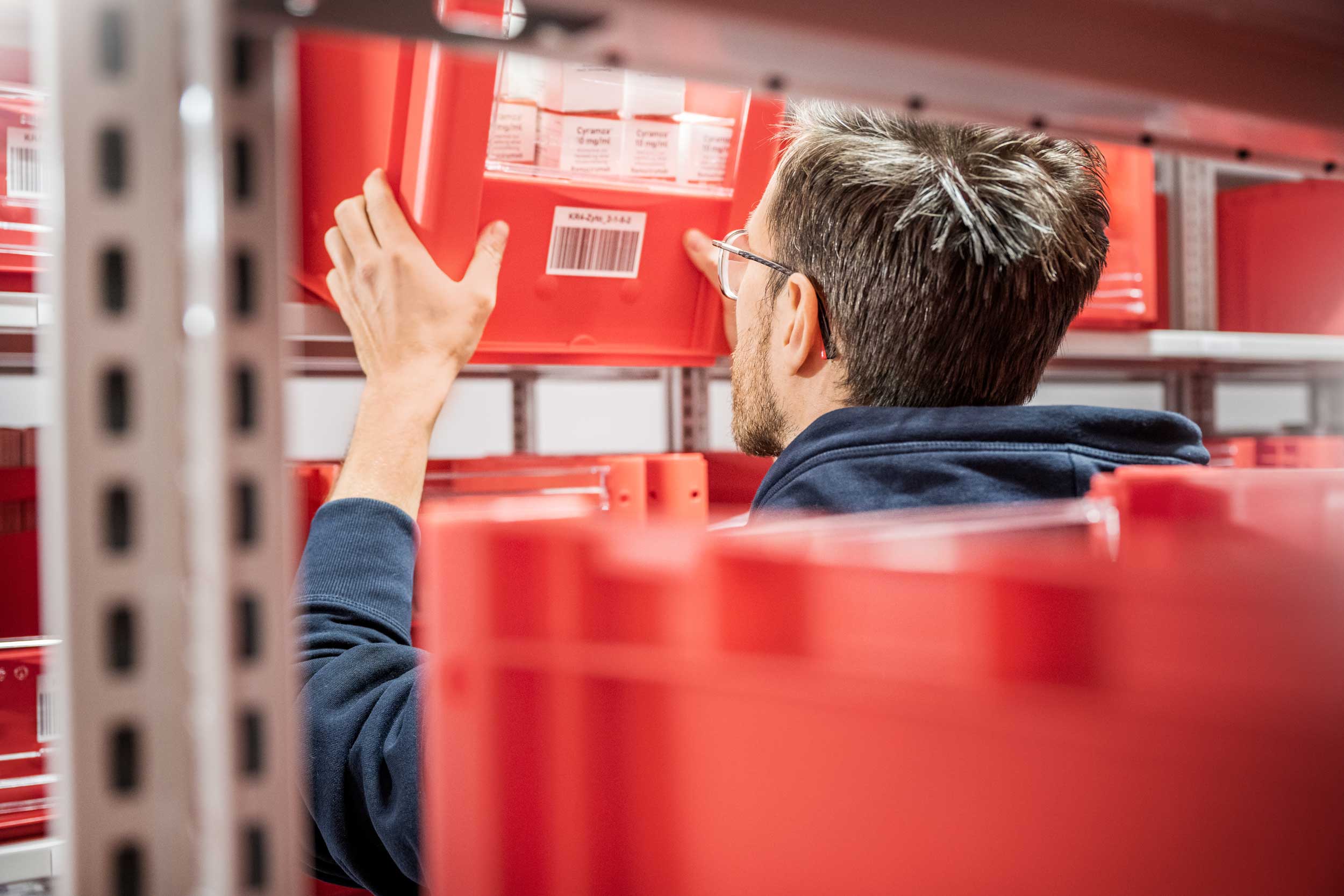 Eine Person greift in einem Lagerregal nach einer roten Box, die mit Medikamenten bestückt ist.